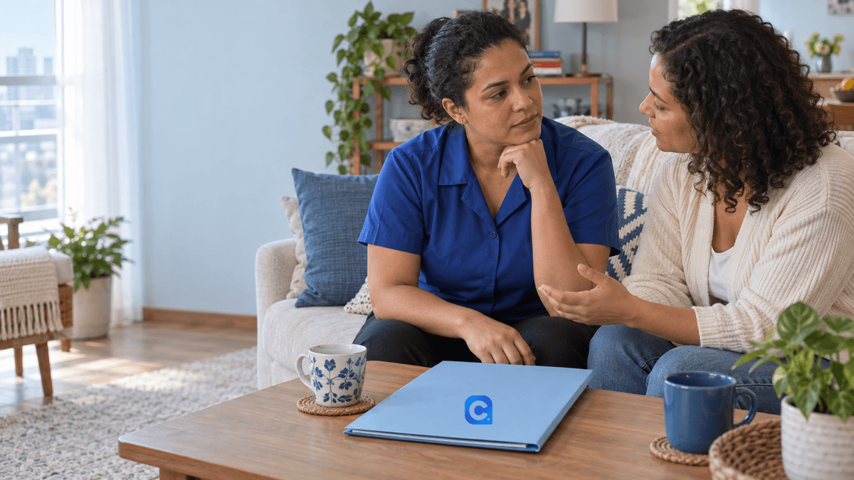 Duas mulheres adultas conversam em casa, ao lado de uma pasta azul sobre a mesa.