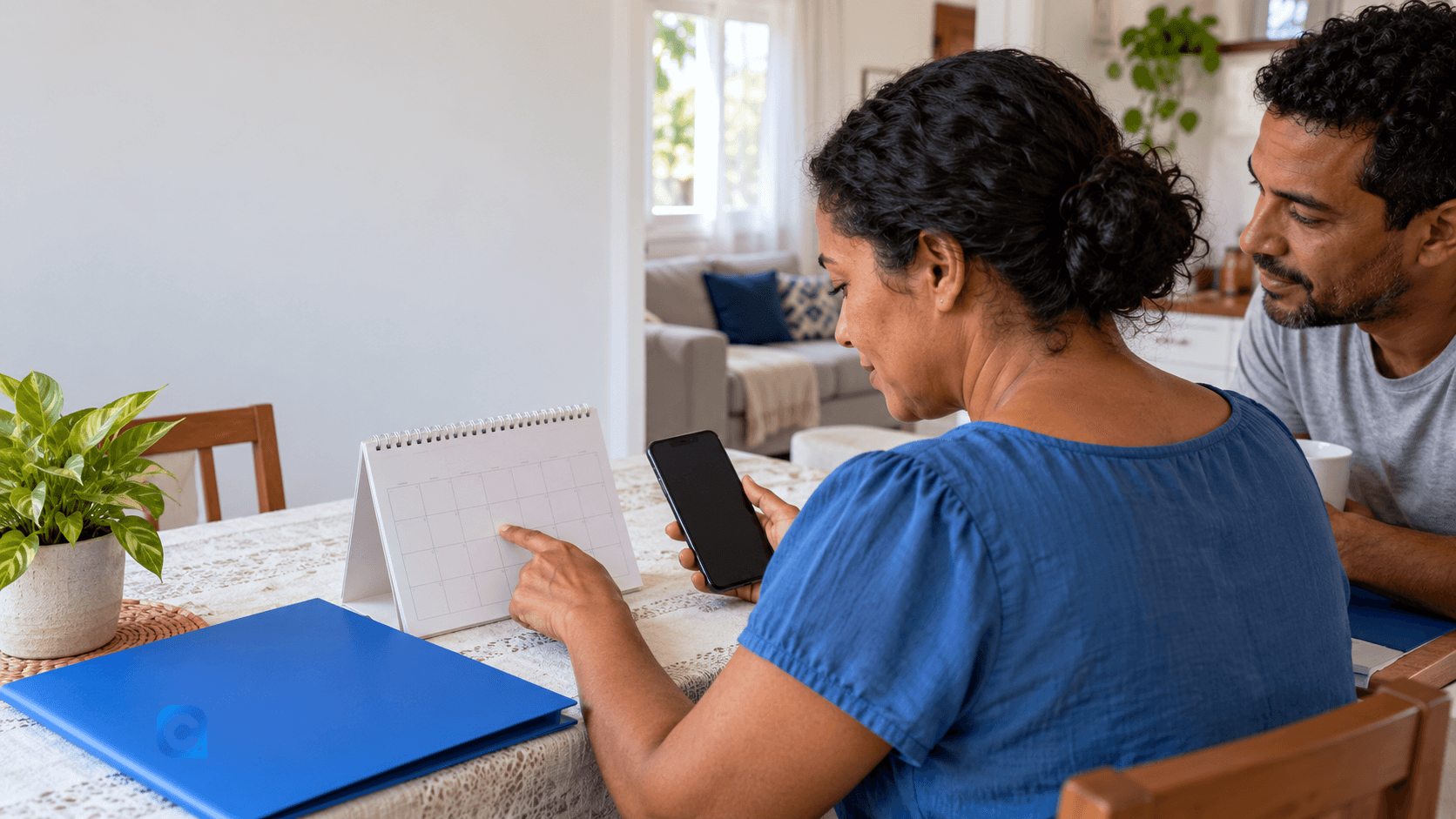 Mulher adulta consulta um calendário e um celular em casa, acompanhada por outro adulto.